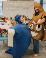 Two people in costumes, one as a roll of toilet paper and one as the poop emoji, smile and pose indoors.