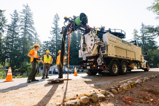Three workers in personal protective equipment stand next to a truck with a large hose extending into an open maintenance hole to access a sewer pipe.