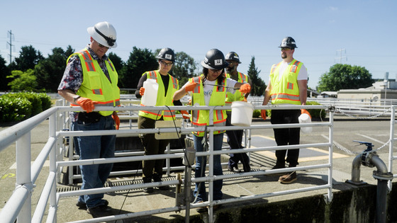 A group of people in personal protective equipment stand behind a railing as they collect wastewater for testing.