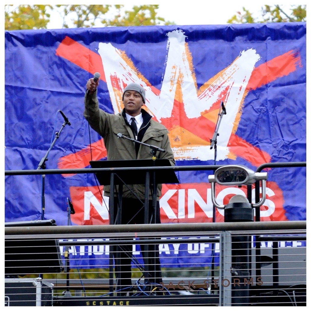 CM Zahilay speaking at the No Kings Rally, holding a microphone up. The No Kings rally logo is in the background.