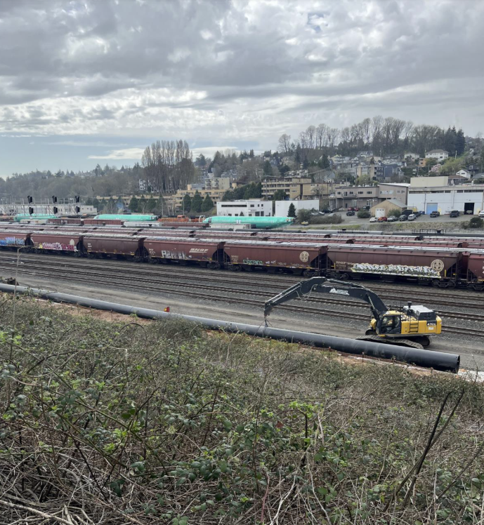 A long black pipe being lifted by an excavator near a train yard.