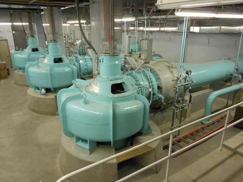 Interior of a wastewater pump station showing turquoise centrifugal pumps connected to heavy-duty pipes and instrumentation.