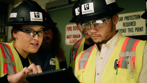 OIT participants in black hard hats, safety glasses, and yellow protective vests work inside an industrial facility.