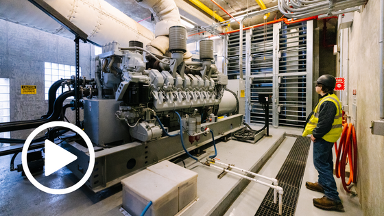 Wastewater operator wearing a safety vest stands beside a large backup generator inside a treatment plant power room.