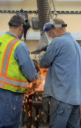 Two workers wearing safety gear grind and weld metal, sending sparks as they repair sewer pipe equipment.