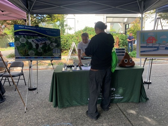 Two people stand outside on either side of a table with project materials.