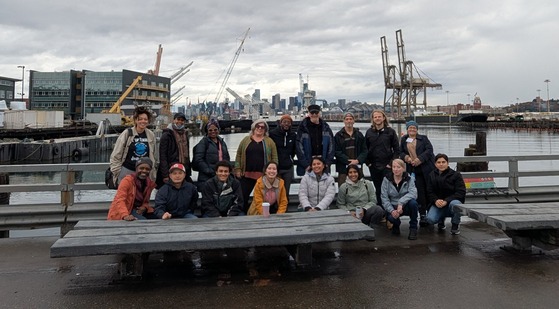 17 personas posando en una pasarela pavimentada, con el río Duwamish al fondo.