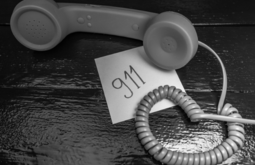 A vintage telephone handset rests on a dark wooden surface next to a piece of paper with "911" written on it and a coiled cord.