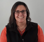 A person with dark hair, glasses, wearing an orange shirt and a black vest, smiles at the camera. They are wearing a peace sign necklace.