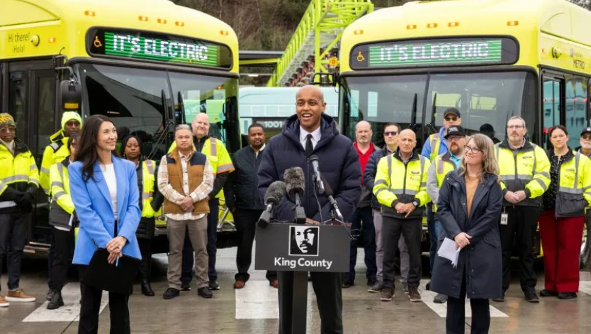 Two electric buses behind Metro employees lined up at press conference with Girmay Zahilay speaking at podium 
