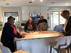 A group of four women stand around a kitchen island cutting fabric patterns on a large cutting mat