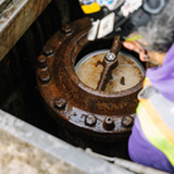 Worker in a purple sweatshirt and safety vest inspects a rusted air valve inside a concrete vault.