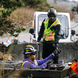 Two workers in safety gear repair a valve inside a concrete vault as one hands a tool down from ground level.