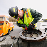 Worker in hard hat and safety vest uses a power tool to clean a rusted valve flange on a concrete ledge.