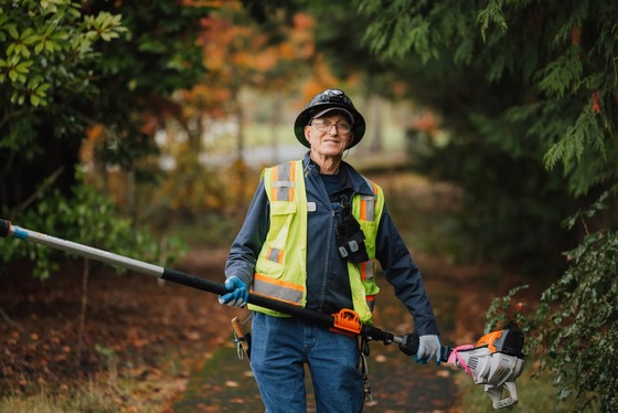 Worker in safety vest and hard hat holding a pole trimmer on a tree-lined path with fall foliage in the background.
