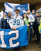 Group of Seahawks fans in jerseys and beanies holding team flags and cheering together outside a building.