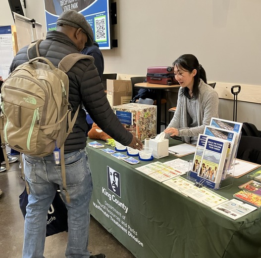 Two adults stand on either side of a table covered in a green tablecloth. On the table, there are informational materials and activities. 
