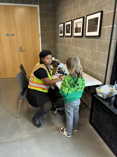 An adult wearing a yellow vest sits on a chair next to a microscope. She is looking at a child who is facing her wearing a green sweatshirt. 
