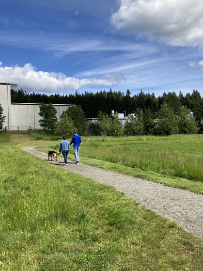Two adults in blue shirts walk their leashed dog on a gravel trail. A concrete building is behind a fence in the background.