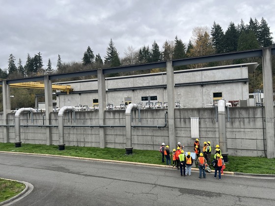 A tour group in bright vests and yellow hard hats stand next to large pipes and an example membrane filter in an outdoor area at the plant.
