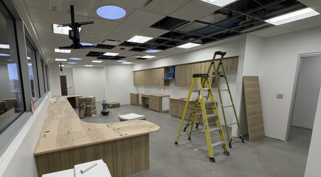 Room with cabinets hanging and unassembled pieces leaning against wall. Ceiling shows missing panels. Two ladders stand in center of the room.