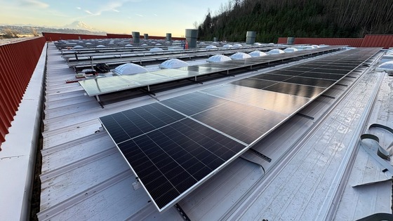 A metal roof outfitted with an array of solar panels. In the distance are trees and Mt. Rainier on a sunny day. 