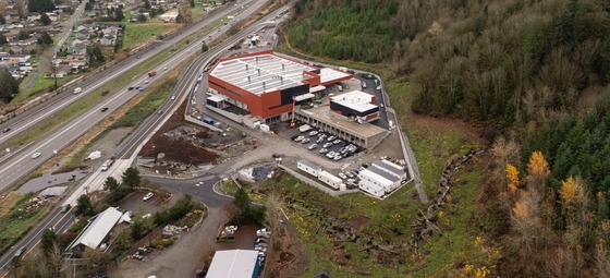 Aerial view of a large industrial complex with a red and black building, a parking lot, and a highway nearby, surrounded by trees.
