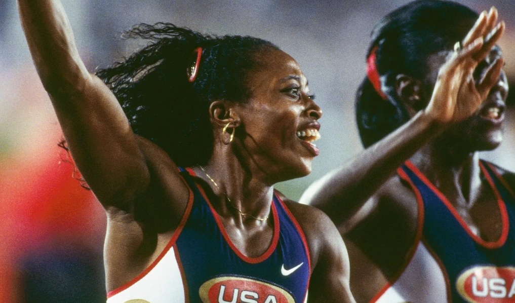 Two female track athletes in USA uniforms celebrating a victory, with one raising her arm in triumph.