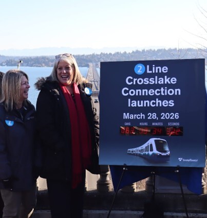 Two smiling people stand next to a sign announcing the March 28 opening of the Crosslake Connection