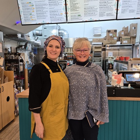 Two women stand and smile in front of cafe cashier counter with a digital menu above them.