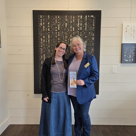 Two women stand and smile in front of a painting mounted on a white wall.
