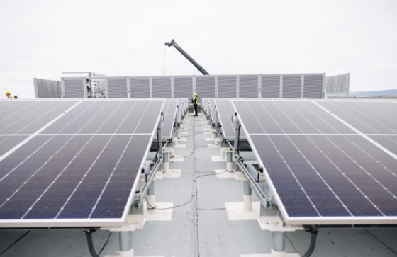 A worker walking between rows of rooftop solar panels, with additional equipment and a crane visible in the background under an overcast sky.