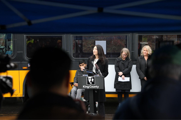 Councilmember Fain in front of a bus and three individuals giving remarks by a podium