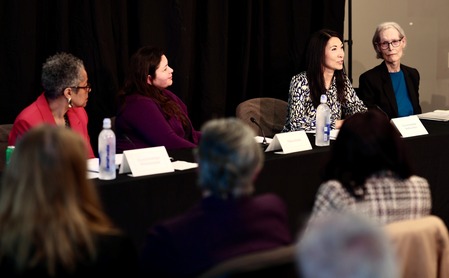 CM. Fain sitting at a table speaking into a mic with three other women next to her in front of a black curtain background