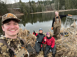 Four young people wearing waders and smiling on the bank of a pond next to a round wire cage.