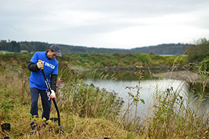  A man standing on a grassy riverbank using a shovel and surrounded by potted plants