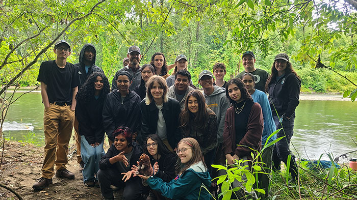 A group of about 20 young people alongside a river with a small stuffed animal in the shape of a beaver