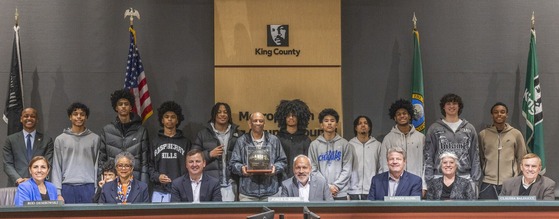 King County Councilmembers with the Rainier Beach Vikings Boys Basketball Team & their coach at the center holding a trophy in the Council Chambers.