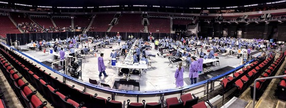 A photo of people in scrubs along rows of tables on the floor of an arena surrounded by tiers of red seats