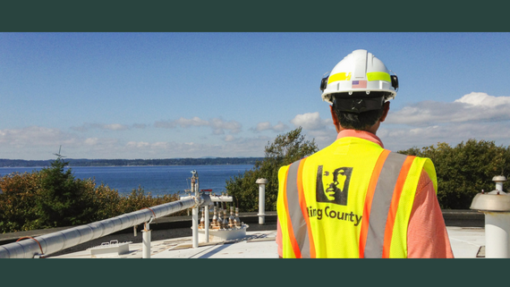 Back of person in high visibility neon yellow and orange vest with King County logo and a hard hat with mountains and blue sky behind.