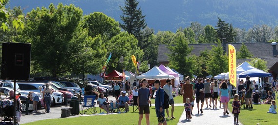 A crowd of people at a farmers' market in a park on a sunny day. There are many colorful tents and tall trees full of green leaves behind.