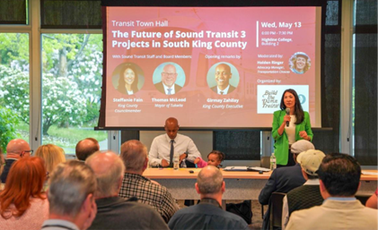 Steffanie speaking in front of a projector with dozens of individuals in audience and the executive sitting at the table with his daughter