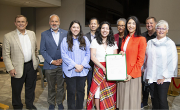 Steffanie holding up a proclamation alongside eight other individuals in council chambers