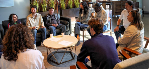 Steffanie sitting down with a coffee cup alongside eight individuals in a lobby with a table in the center