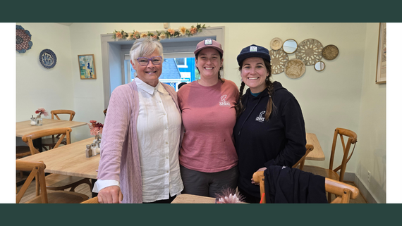 Three women stand, smiling, in a coffeeshop.