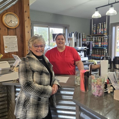 A woman stands, smiling, in a black and white coat and a woman in a red t-shirt standing behind a countertop.