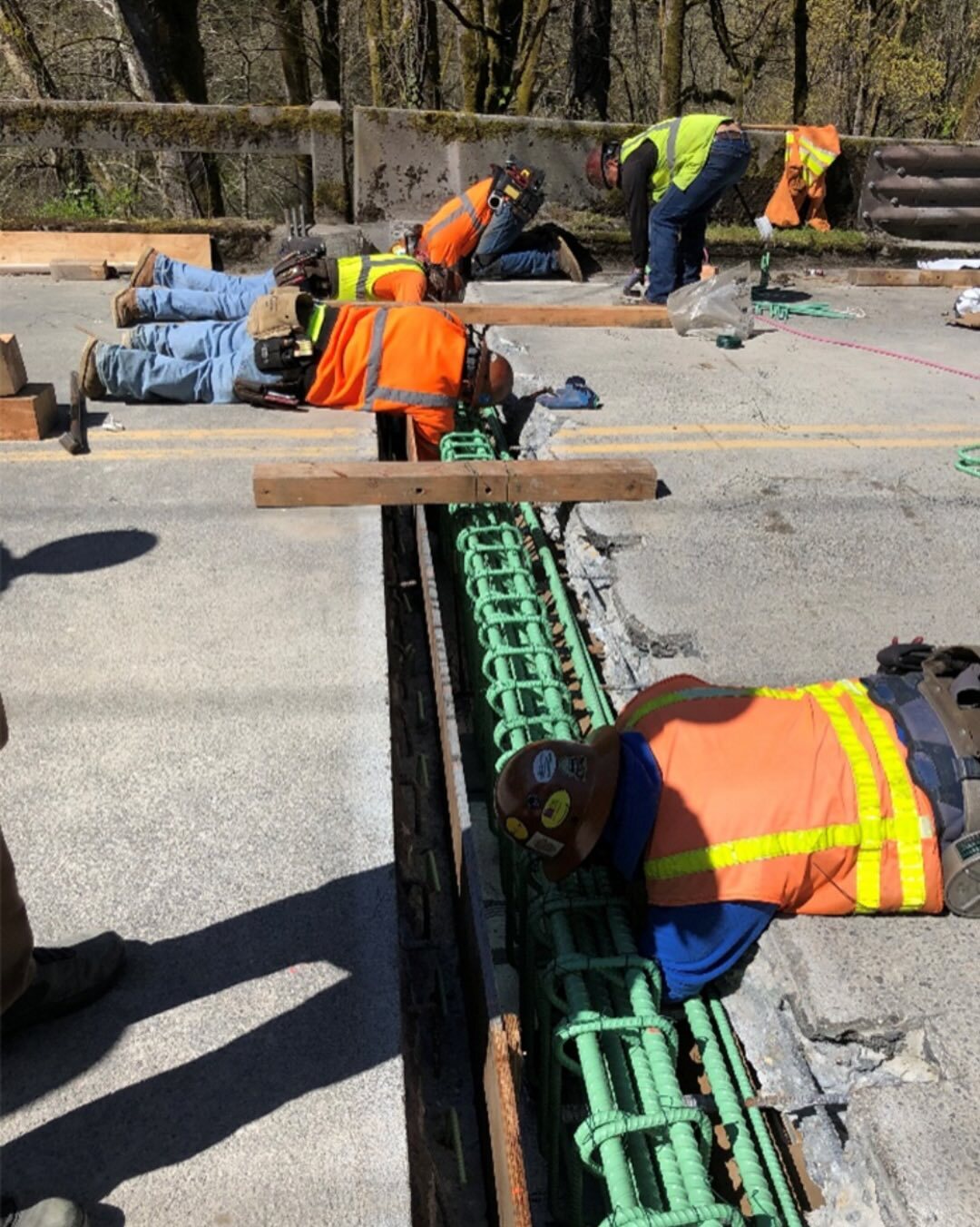 Five people in orange and yellow safety vests lay on a road fixing a green joint in a bridge
