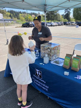 WTD staff stand behind their table at a community event and helps a child compares how different items dissolve in water.
