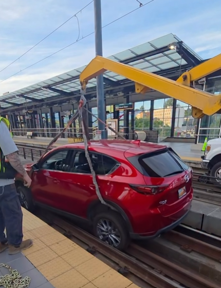 A red car being hoisted off light rail tracks by a yellow crane with a man in a yellow safety vest in the foreground