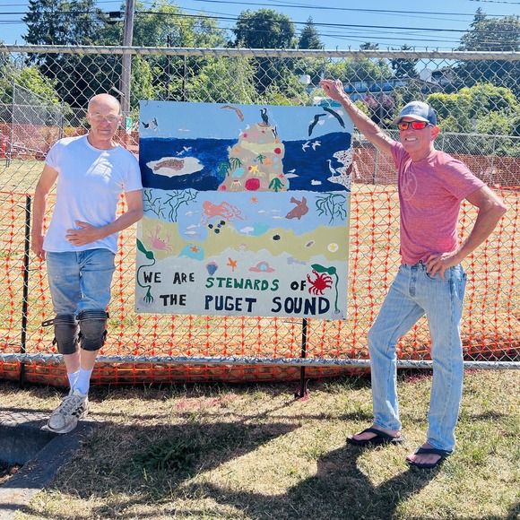 Two men stand and pose around a piece of art on a construction fence.